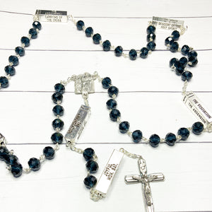 A deep blue mysteries rosary lays on a white washed wooden background
