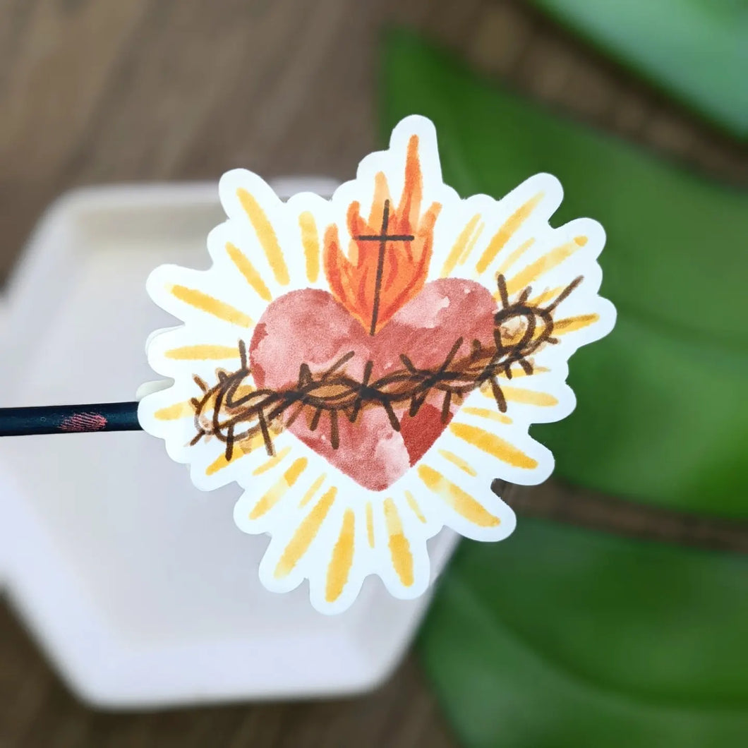 A watercolor style sacred heart vinyl sticker is held abover a blurred white and green background