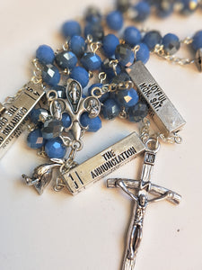 A close up of A light blue and silver catholic rosary that has silver bars that list the mysteries of the Rosary on each bar which lays against a white plate 