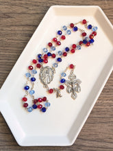 Load image into Gallery viewer, A rosary featuring shades of red and blue with a divine mercy centerpiece lays in a white tray with a wooden background