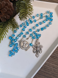 An aqua blue rosary with a mother and child centerpiece lays in a white tray with pine needles and a pine cone next to it. 