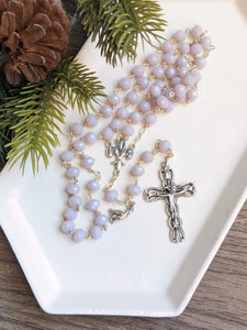 A lilac colored rosary sets in a white tray with a pine branch next to it