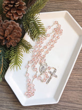 Load image into Gallery viewer, A clear pink glass bead rosary with a St Agatha centerpiece lays in a white tray with a pine branch next to it with a wooden background