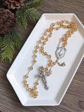 Load image into Gallery viewer, A golden glass bead rosary with a fatima centerpiece lays in a white tray with a pine branch next to it on a wooden background