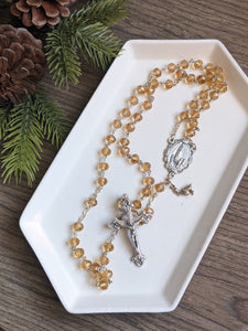 A golden glass bead rosary with a fatima centerpiece lays in a white tray with a pine branch next to it on a wooden background
