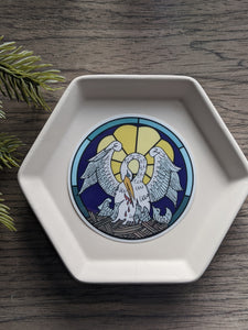 A pelican in her piety round circle sticker sits on a white tray. The background is wood and a decorative plant 