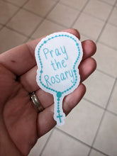 Load image into Gallery viewer, A teal rosary shaped sticker with the words pray the rosary in the center is being held in a hand