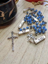 Load image into Gallery viewer, A light blue and silver catholic rosary that has silver bars that list themysteries of the Rosary on each bar lays on a wooden board with a black and gold coffee cup next to it. 
