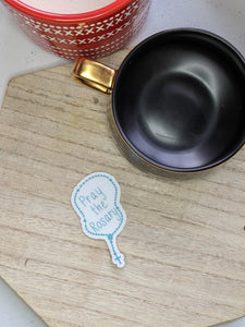 The osary shaped sticker with the words pray the rosary is sitting on a wooded tray next to a black coffeemug and a red candle
