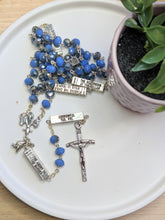 Load image into Gallery viewer, A light blue and silver catholic rosary that has silver bars that list themysteries of the Rosary on each bar laus against a white plate with a small fake plant next to it. 