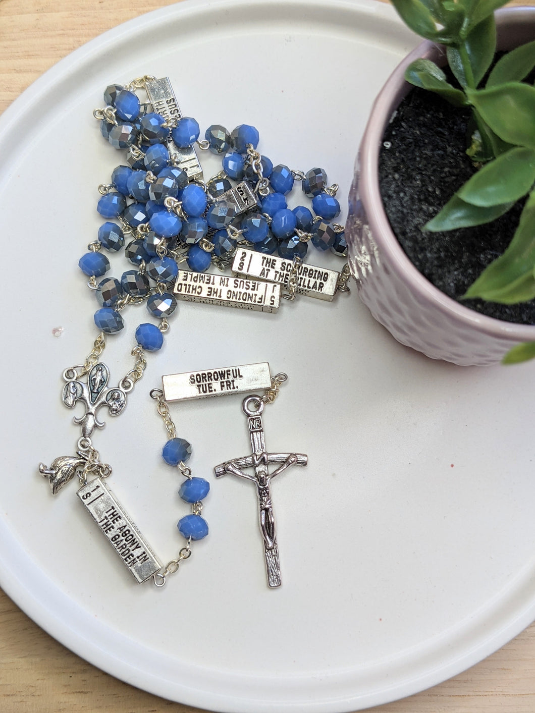 A light blue and silver catholic rosary that has silver bars that list themysteries of the Rosary on each bar laus against a white plate with a small fake plant next to it. 