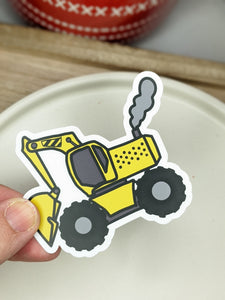 A cartoon excavator sticker is held abover a white dish with a wooden tray and a red vase in the background. 