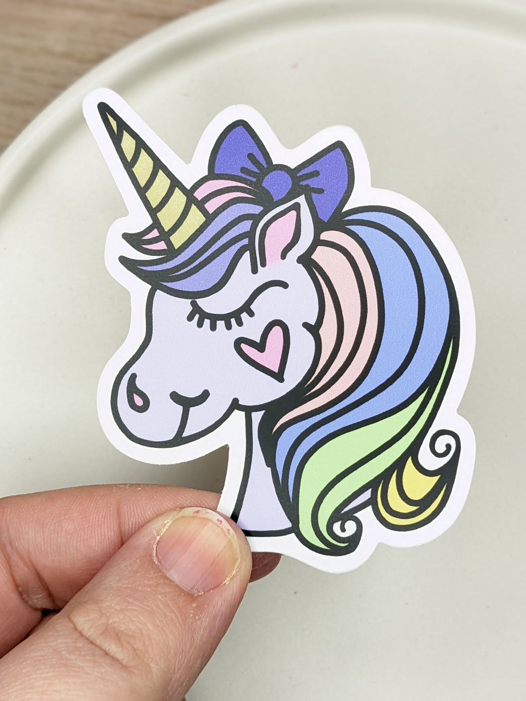 A cartoon style pastel unicorn sticker is being held over a white dish with a wooden background