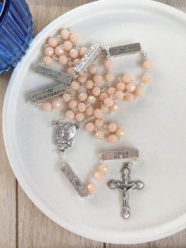 A peach mysteries rosary lays in a white tray on a wooden background with a blue cup next to it. 