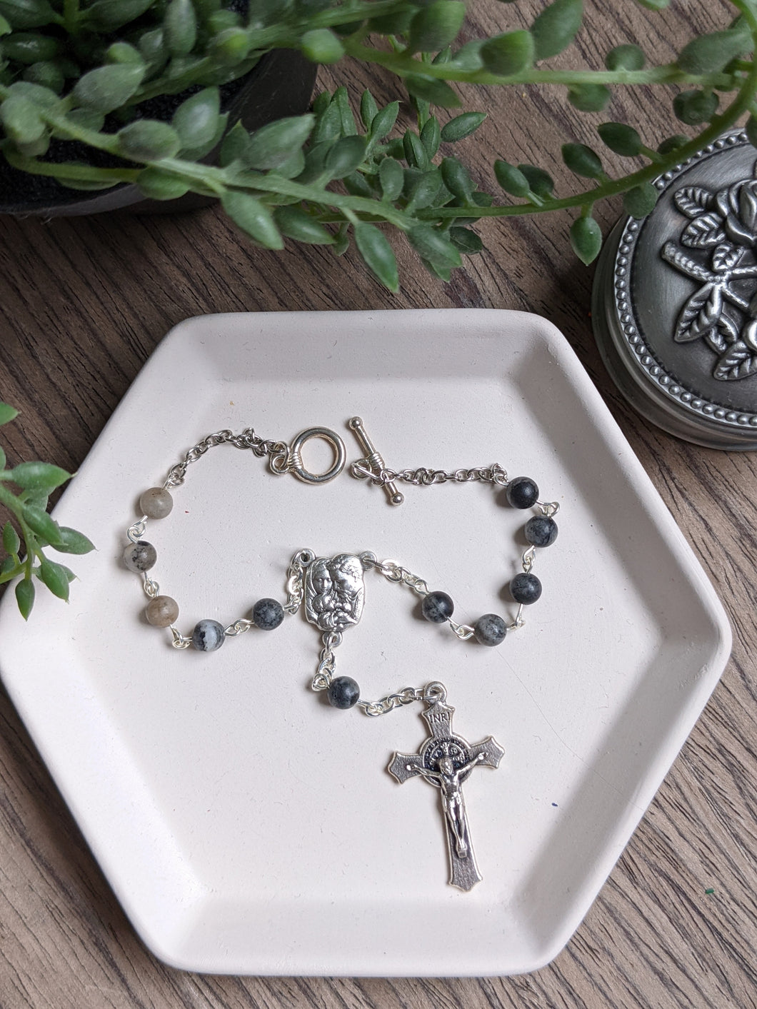A small jasper stone car rosary featuring a holy family centerpiece and st benedict crucifix lays on a white tray. The background is wood with a plant and a rosary holder