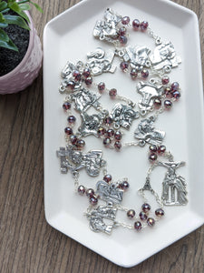 A large stations of the cross chaplet lays in a white tray. It features purple crystal glass beads and all 14 stations of the cross depicted on chaplet medals. 