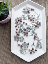 Load image into Gallery viewer, A large stations of the cross chaplet lays in a white tray. It features purple crystal glass beads and all 14 stations of the cross depicted on chaplet medals. The tray lays on a wooden background with a small plant next to it