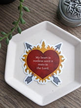 Load image into Gallery viewer, The flaming heart shaped sticker features the words My heart is restless until it rests in you. The sticker rests on a white tray with a wooden background with a decorative ppant and rosary holder in the background