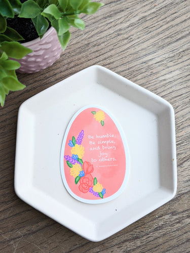 A pink easter egg shaped sticker features flowers and the quote be himble, be simples and bring joy to others. The sticker reats on a white tray with a wooden background and a plant to the left