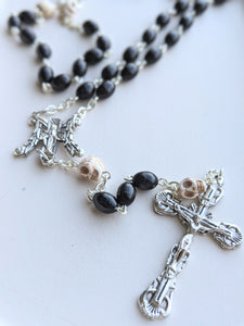 A close up of the black oval wooden shaped bead rosary with skull shaped oir father beads, an ornage M shaped centerpiece for Mary and a skull engraved crucifix
