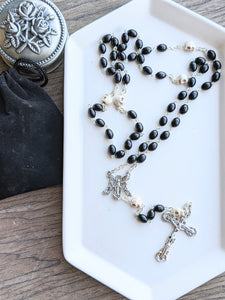 A full sized rosary featuring oval shaped black wooden beads and skull shaped our father beads lays on a white tray. The tray is set on a wooden background with a rosary holder next to it. 