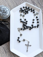 Load image into Gallery viewer, A full sized black crystal glass bead rosary with medal roses for the Our Father beads lays on a white tray. The tray is set on a wooden background with a rosary holder next to it. 