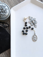 Load image into Gallery viewer, A Black pocket rosary with a skull Our Father bead, a St Joseph medal and an ornate crucifix lays in a white tray. The tray is on a wooden background with a rosary holder next to it 
