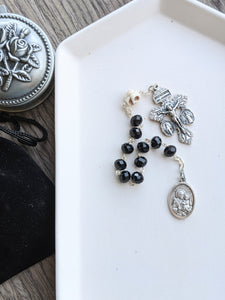 A Black pocket rosary with a skull Our Father bead, a St Joseph medal and an ornate crucifix lays in a white tray. The tray is on a wooden background with a rosary holder next to it 