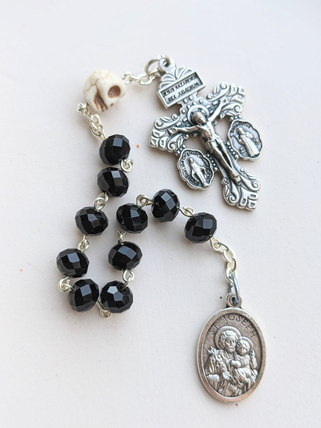 A black glass bead pocket rosary fearuring a skull shaped our father bead, a St Joseph medal and an prnate Pardon of Indulgence crucifix lays on a white background