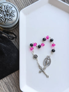 A 10 bead chaple with black and hot pink crystal.glass beads for the hail mary beads and a medal rose bead for the Our Father Bead lays on a white tray. A moraculous medal is attache to one end and a small decorative crucifx is attached to the other aide. The tray lays on a wood background with a rosary holder next to it. 