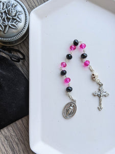 A 10 bead chaple with black and hot pink crystal.glass beads for the hail mary beads and a skull shaped bead for the Our Father Bead lays on a white tray. A miraculous medal is attache to one end and a small decorative crucifx is attached to the other aide. The tray lays on a wood background with a rosary holder next to it. 
