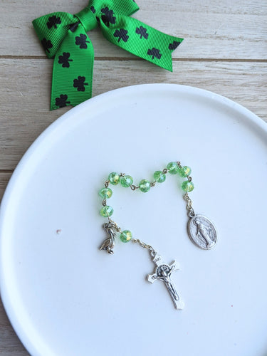 A light green glass pocket rosary with a miraculous medal and a pelican charm sit on a white dish with a wooden background and a green and black shamrock bow for decor next to the dish