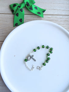 A dark emerald green glass bead rosary bracelet lays on a white dish with a wooden background and a green and black shamrock bow for decor