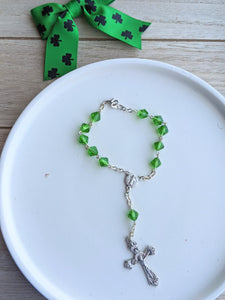 A Peridot green colored glass bead car rosary with a small st Benedict's medal centerpiece and an ornate crucifix lays on a white teay with a wooden background and decorative bow 