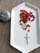 Load image into Gallery viewer, A solid red bead rosary with a dove style goly spirit centerpiece lays against a white tray with a silver rose rosary holder next to it. 