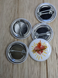 4 button pins lay upside down on a wooden background so you can see the pin style clasp. A single pin is facing up to show the watercolor style sacred heart on a white background. 