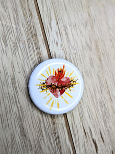 A watercolor style sacred heart is on a white bround button pin. A wood backdrop is behind the pin. 