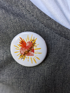 A watercolor style sacred heart is on a white background button pin attached to a grey and white cloth for background. 