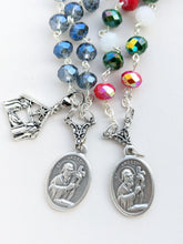 Load image into Gallery viewer, 2 St Andrew Chaplets sit on a white background. The chaplet on the left is a clear and transparent crystal bead with a St Andrew medal and a Nativity Charm. The Right features red and green metallic crystal beads, and a white bead in an alternating pattern as well as a St Andrew medal. 
