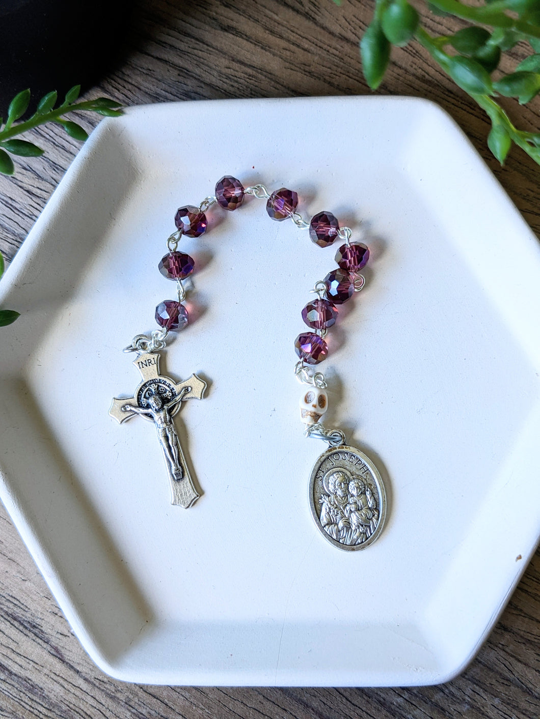 A purple glass bead pocket rosary sits on a white tray with a wooden background and a plant hangs from the top of the frame. 