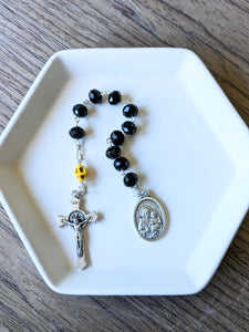A black glass bead pocket rosary with a yellow colored skull for the our father bead sits in a white tray with a wooden background