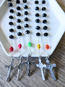 5 black glass bead pocket rosaries featuring a different colored skull all set in a white tray with a wooden background