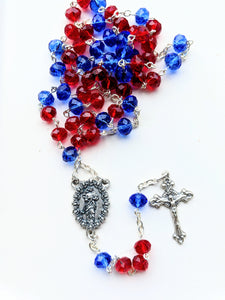 A red and blue glass bead Our Lady Undoer of Knots rosary sits on a white background. 