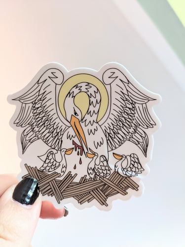 A hand drawn Pelican in her Piety on a white background die cut sticker is held over a white background. 