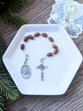 Load image into Gallery viewer, A wooden bead St Padre Pio 9 bead chaplet lays on a white tray with a pelican sticker to the right on a wood and fern background