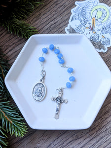 A light blue crystal glass St Padre Pio 9 bead chaplet lays on a white tray with a pelican sticker to the right on a wood and fern background