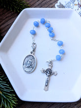 Load image into Gallery viewer, A light blue crystal glass bead St Padre Pio 9 bead chaplet lays on a white tray with a pelican sticker to the right on a wood and fern background