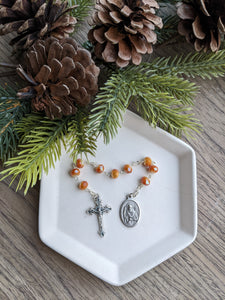 A orange gold St Nicholas Chaplet sits on a white tray with a wooden background and a everygreen branch with pinecones framing the top of the photo