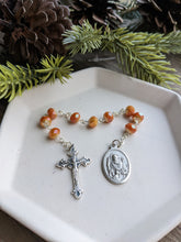 Load image into Gallery viewer, A orange gold St Nicholas Chaplet sits on a white tray with a wooden background and a everygreen branch with pinecones framing the top of the photo