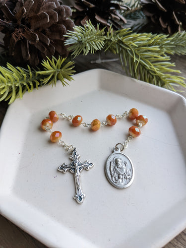 A orange gold St Nicholas Chaplet sits on a white tray with a wooden background and a everygreen branch with pinecones framing the top of the photo